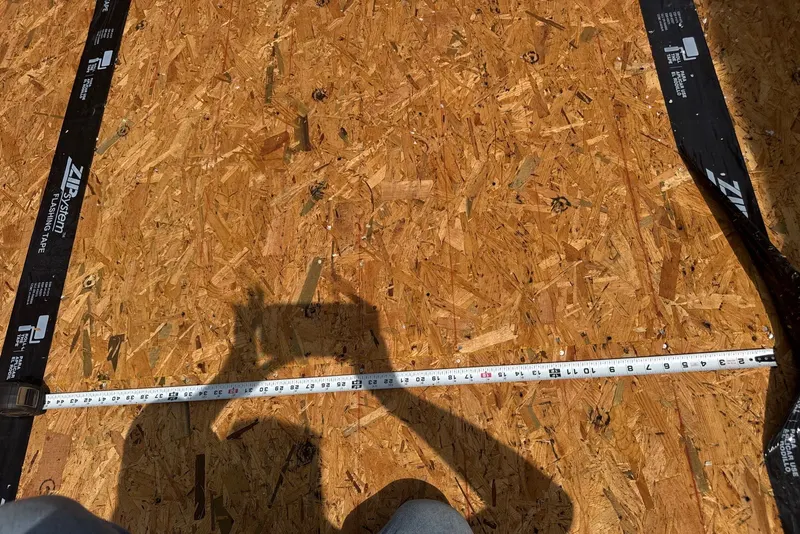 Close-up of black flashing tape sealing a seam on an OSB roof deck, with a tape measure